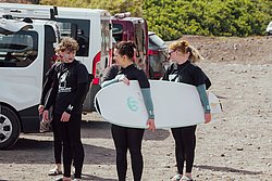 Three young surfers in wetsuits holding surfboards near vans at a beach parking lot with green hills behind them.