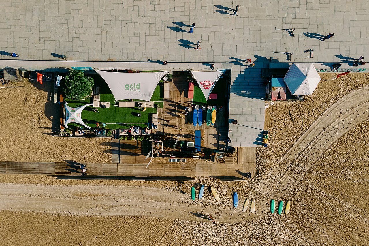 Aerial view of the Onda Pura surf school at Matosinhos beach, with surfboards, shaded seating areas, and people walking along the beachfront promenade.