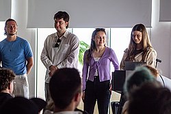 Three young professionals presenting to an audience in a modern office with bright windows and plants