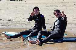 Two young surfers in wetsuits sitting on surfboards in shallow beach water, smiling and waving at camera