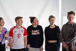 Three young people standing against a white background, wearing casual clothing including a sports jersey, graphic t-shirt, and hoodie.