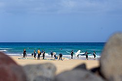 Group of surfers in wetsuits with surfboards on a sandy beach with turquoise ocean and clear blue sky