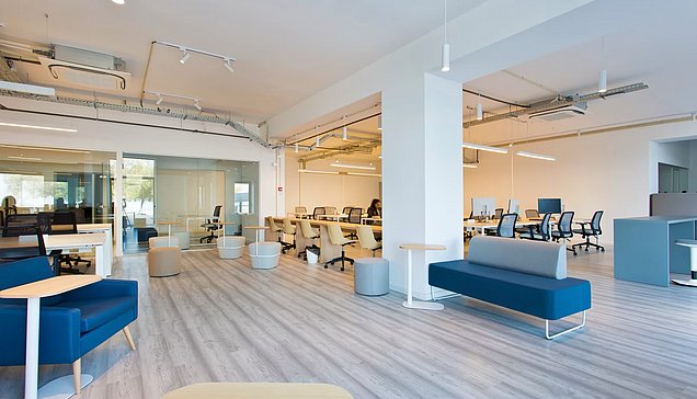 Bright, open coworking space at Edifício Transparente in Porto, featuring desks, office chairs, glass meeting rooms, and blue lounge seating.