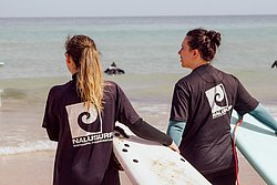 Two female surfers in Nalusurfe wetsuits holding surfboards on beach, ocean in background