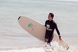 Surfer in black wetsuit carrying beige surfboard walking out of ocean waves