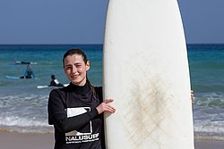 Young surfer in wetsuit holding surfboard at beach with ocean and blue sky in background