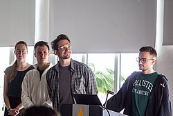 Three men at a podium during a presentation in a bright modern room with large windows and plants visible outside.
