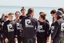 Group of young surfers wearing Nalusurf Surfschool Fuerteventura black shirts standing together on a beach