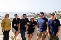 Five people smiling at camera on coastal terrace with the sea and white buildings in background