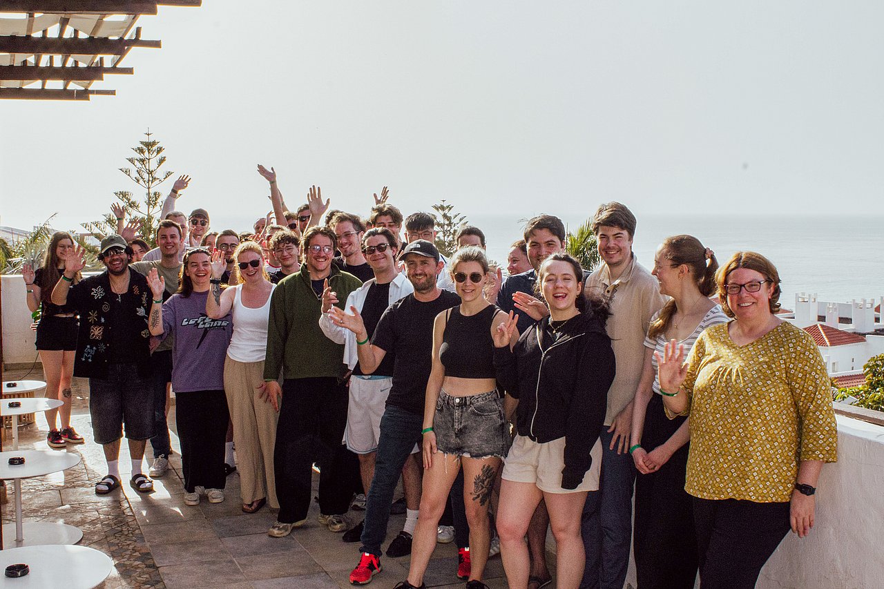 Large group of smiling young people posing together outdoors with raised hands against a coastal sky backdrop