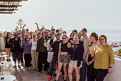 Large group of smiling young people posing together outdoors with raised hands against a coastal sky backdrop